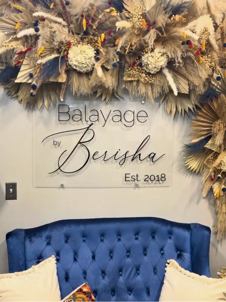 Sign on wall reads "Balayage by Berisha Est. 2018" above a blue tufted sofa and floral decor