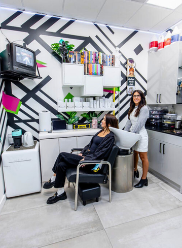 Two women in a modern room with a TV, plants, and shelves, one sitting on a chair