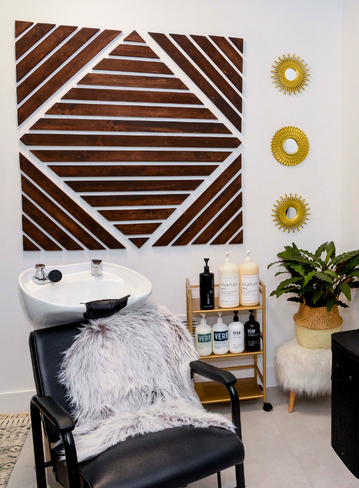 A salon chair with a fur throw, sink, shelves of products, and decorative wall panels