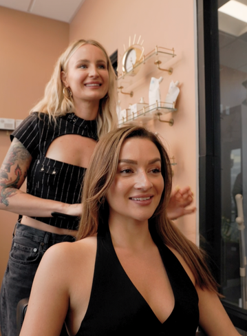A stylist stands behind a woman with long hair, both smiling in a salon setting