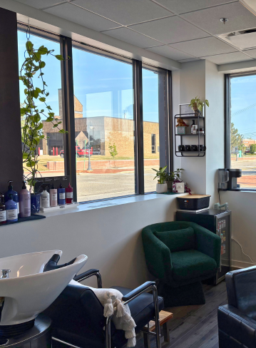 Interior of a salon with windows, plants, and a black shampoo bowl. Seating and shelves visible