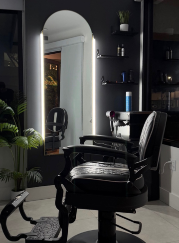 A black salon chair is positioned near a lit mirror and shelves with products