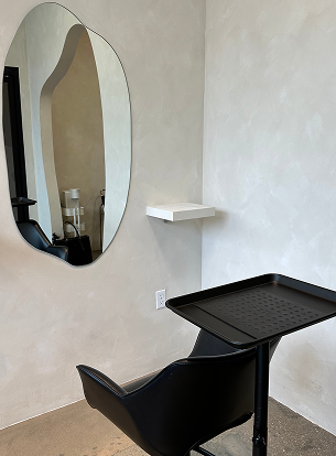 A corner with a black chair, small shelf, and an oval mirror on a textured wall