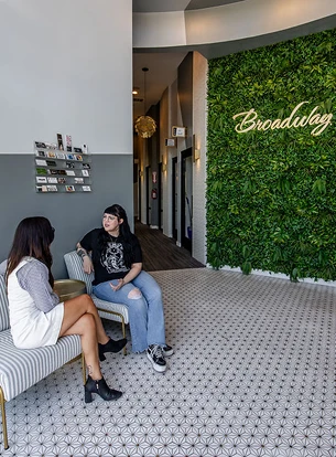 Two women sit on chairs in a modern lobby with green wall decor and patterned flooring