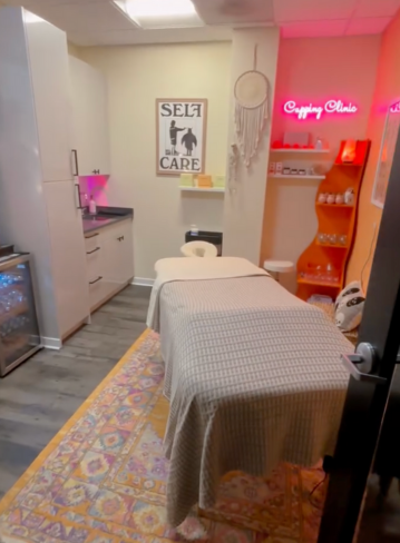 A small treatment room with a massage table, shelves, and a neon sign on the wall